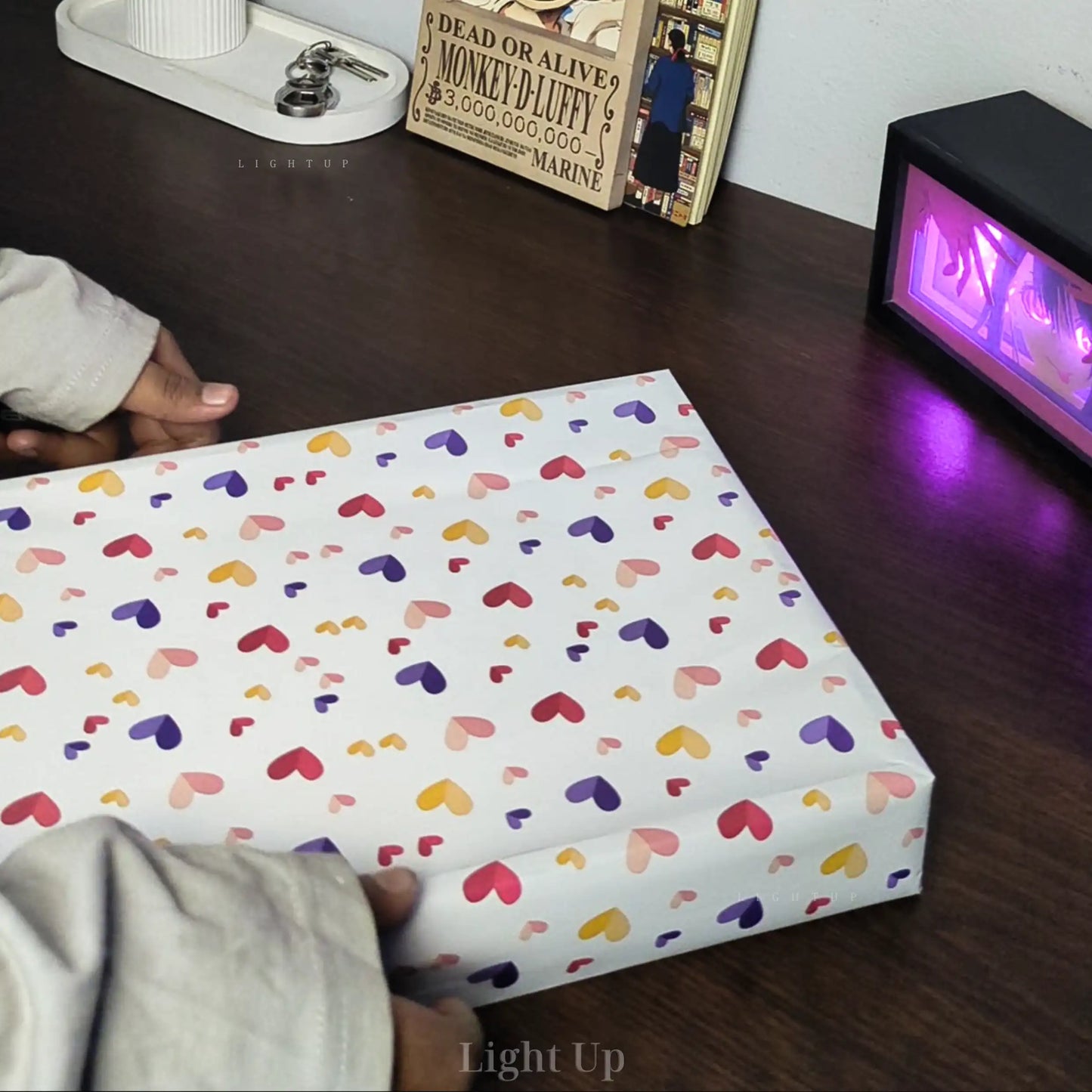 Box with colorful heart pattern on a wooden surface with a person's hand and a clock in the background.